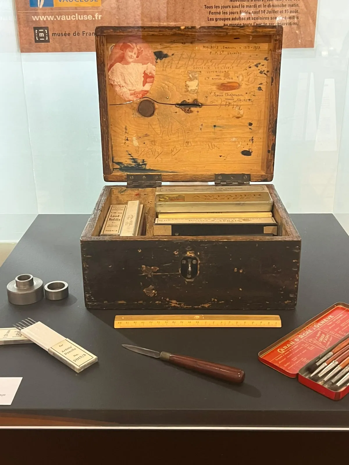 Vue de face sur un coffret bois contenant divers ustensiles rangés dans des compartiments, une photo figure à l'intérieur du couvercle dans le coin, en haut à gauche