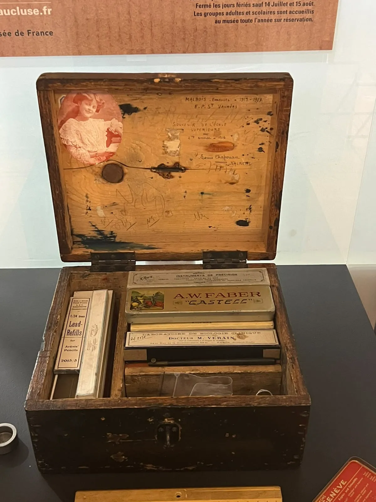 Vue de face sur un coffret bois contenant divers ustensiles rangés dans des compartiments, une photo figure à l'intérieur du couvercle dans le coin, en haut à gauche