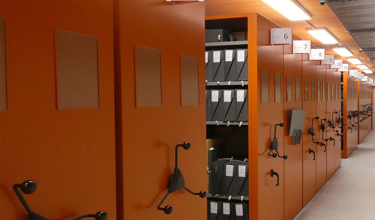Vue sur des compactus métalliques de couleur orange ; sur les tablettes, des boîtes en carton de couleur noires sur lesquelles figurent des références
