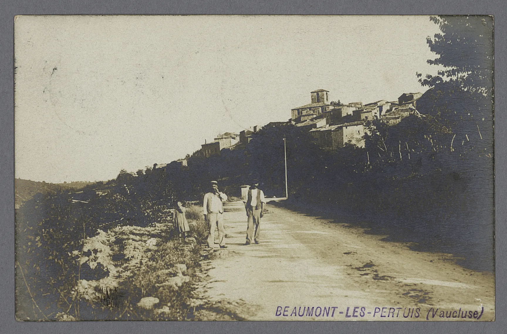 Carte postale en noir et blanc représentant 2 hommes et un enfant sur une route. Au loin, un village