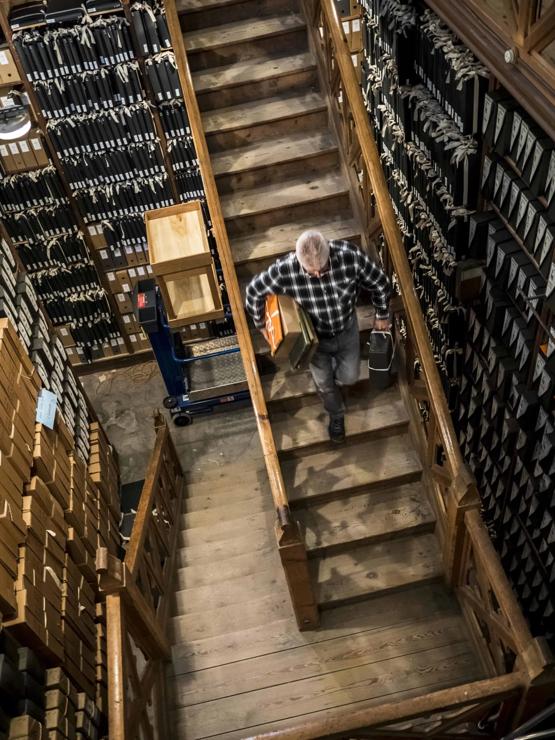 Dans un salle qui semble immense, équipée de rayonnages bois sur lesquels reposent des cartons, un homme descend un escalier de bois, une boîte sous le bras.