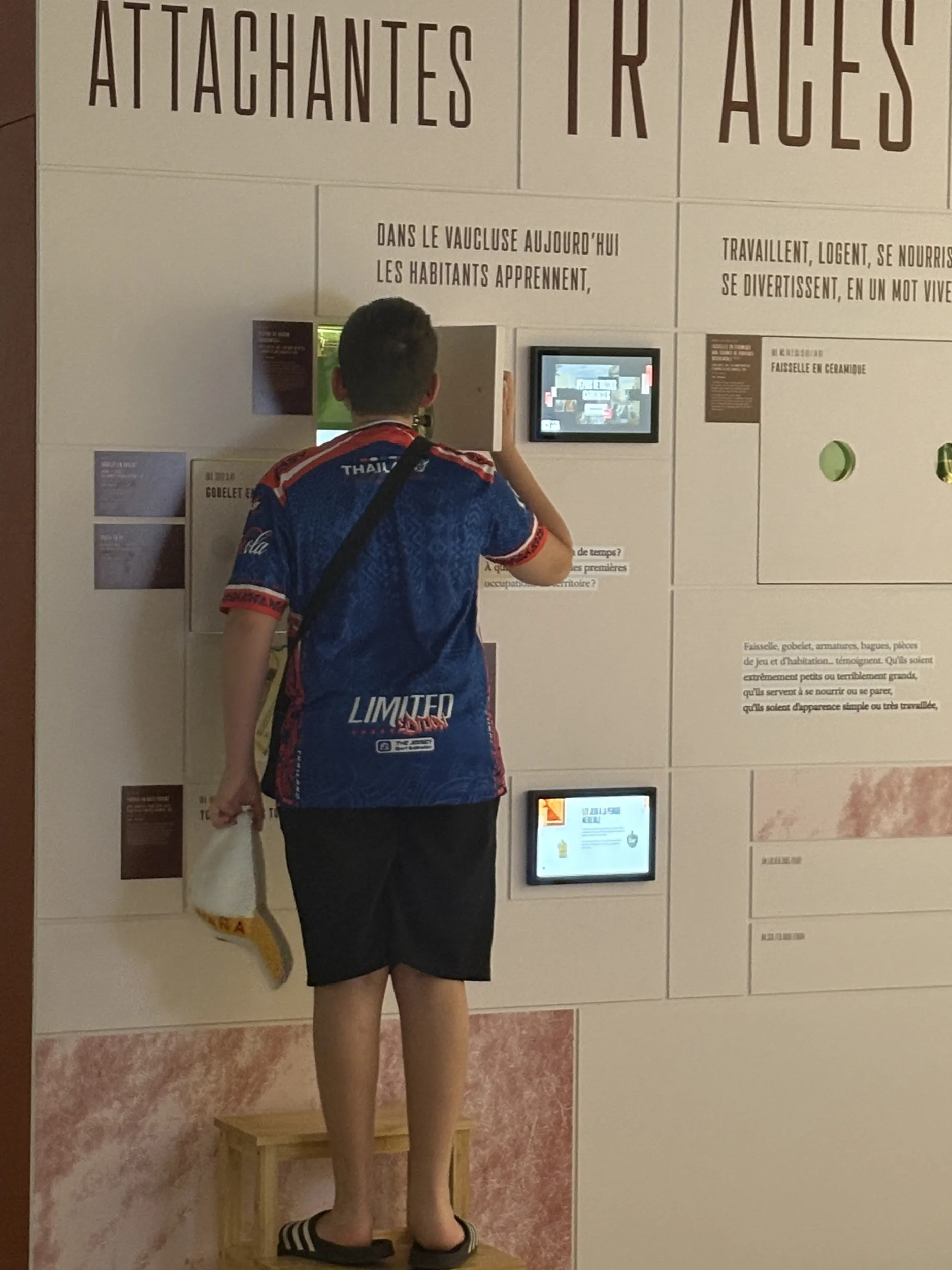 Plan d'ensemble sur un jeune visiteur juché sur un petit escabeau qui ouvre une petite porte pour regarder un objet éclairé