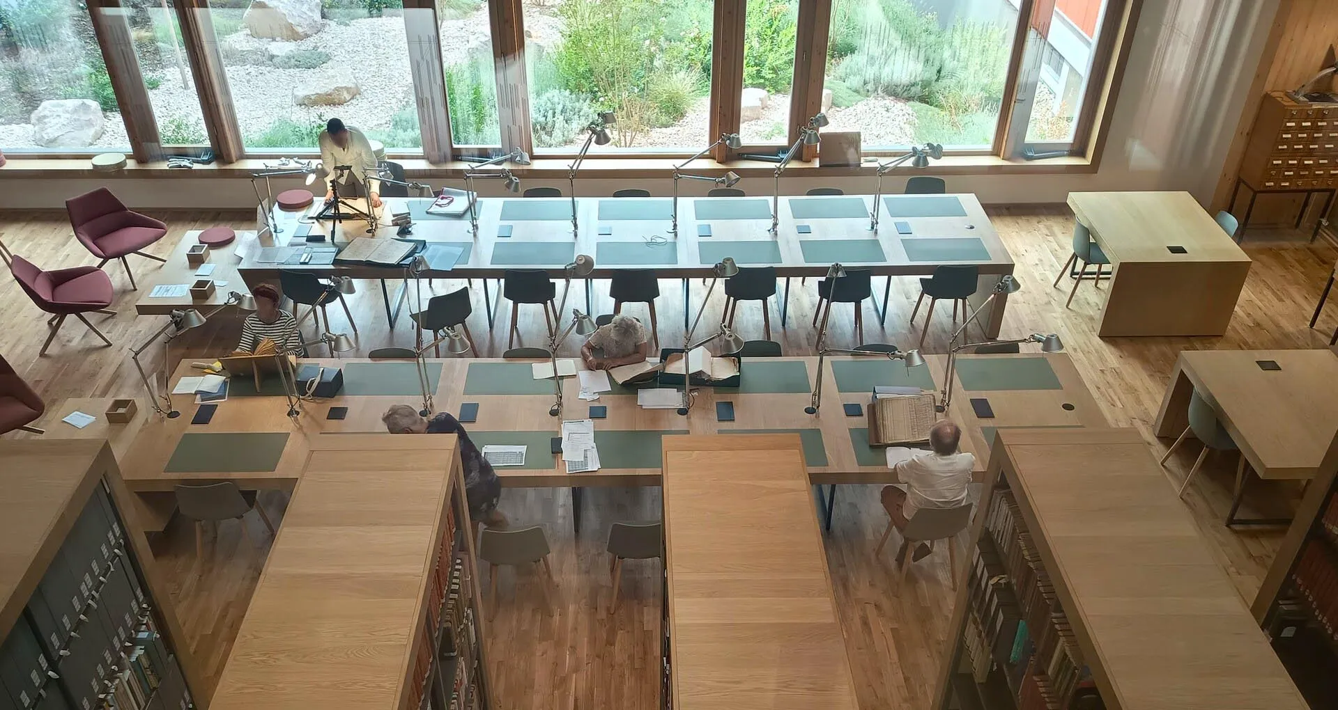 Dans une grande salle équipée de mobilier bois : rayonnages de bibliothèque, 2 grandes tables de travail, baies vitrées en fond, des chercheurs consultent des documents