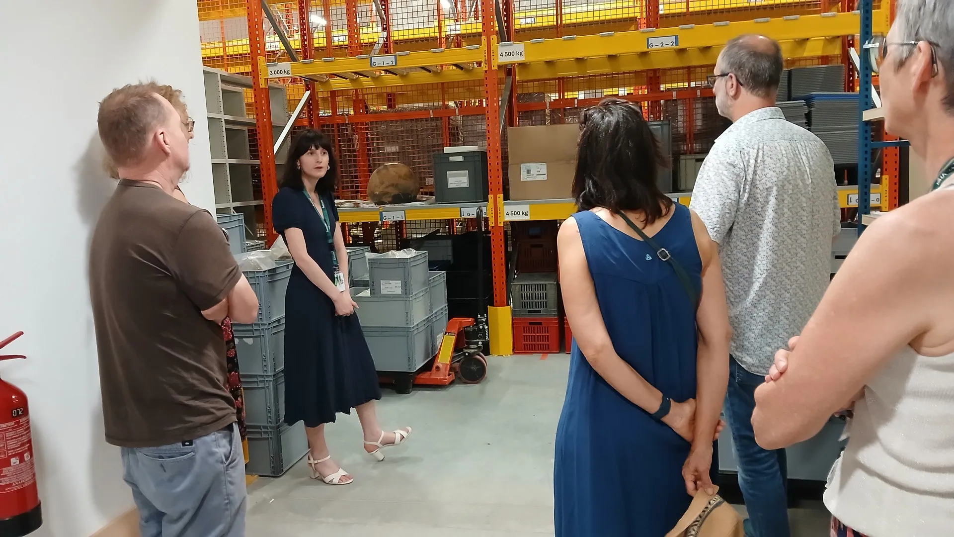 Dans une salle composée de grands racks de couleur jaune et orange sur lesquels reposents des objets et des caisses, une médiatrice présente les missions à un groupe de 5 personnes