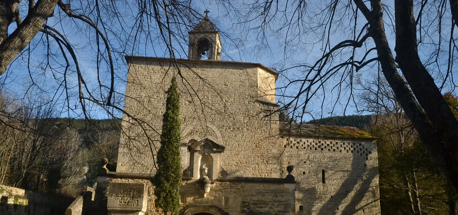 Photographie couleur montrant la chapelle un jour d'hiver. En détail, de gauche à droite, on voit le pigeonnier, la vierge Marie dans une niche, les escaliers montant à la terrasse et le clocher ; des branches d'arbres nues au premier plan
