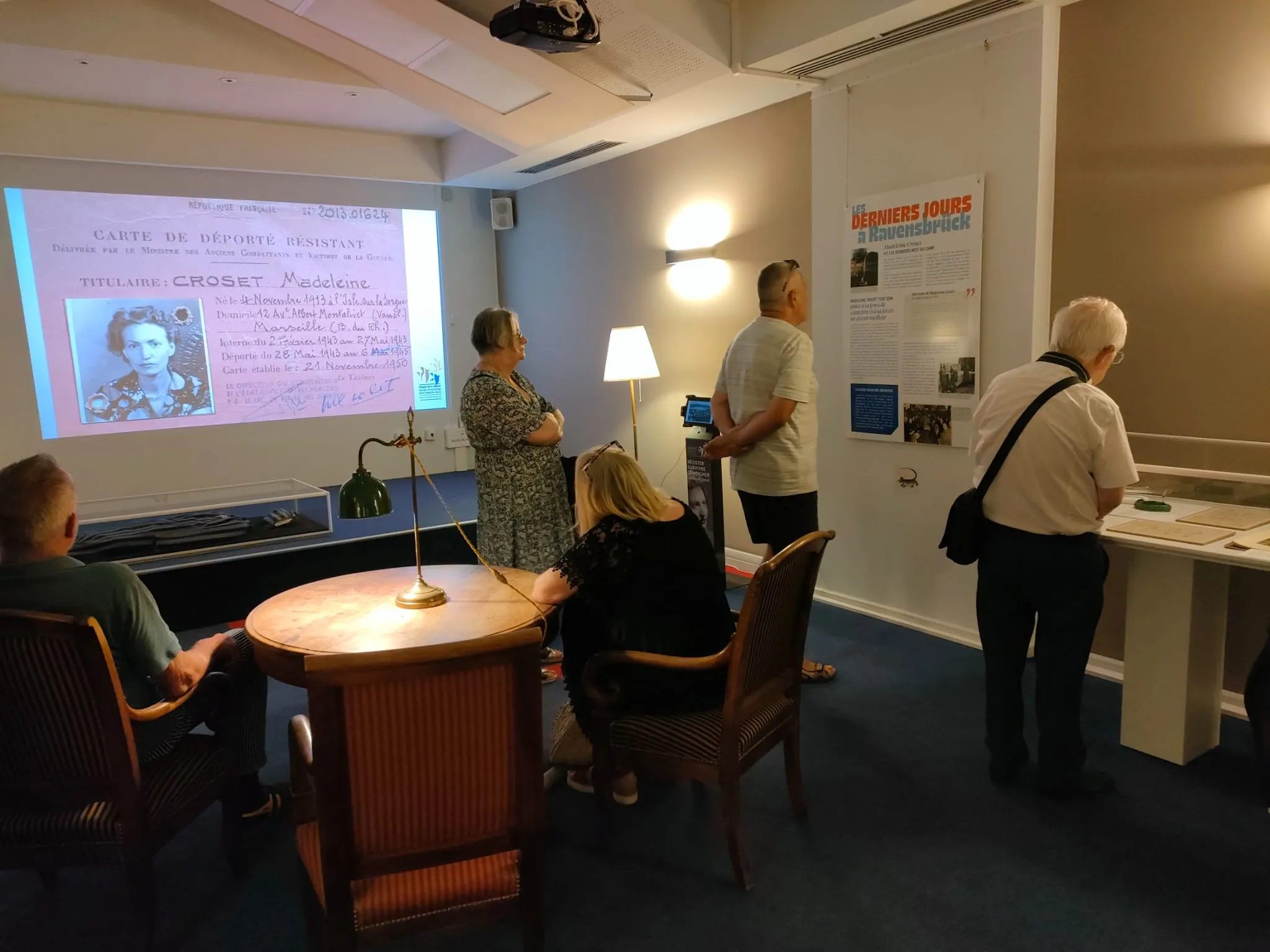 Des visiteurs contemplent les vitrines et panneaux d'une exposition ; en fond, un grand écran sur lequel figure la carte [rose] de déporté résistant de Madeleine Croset