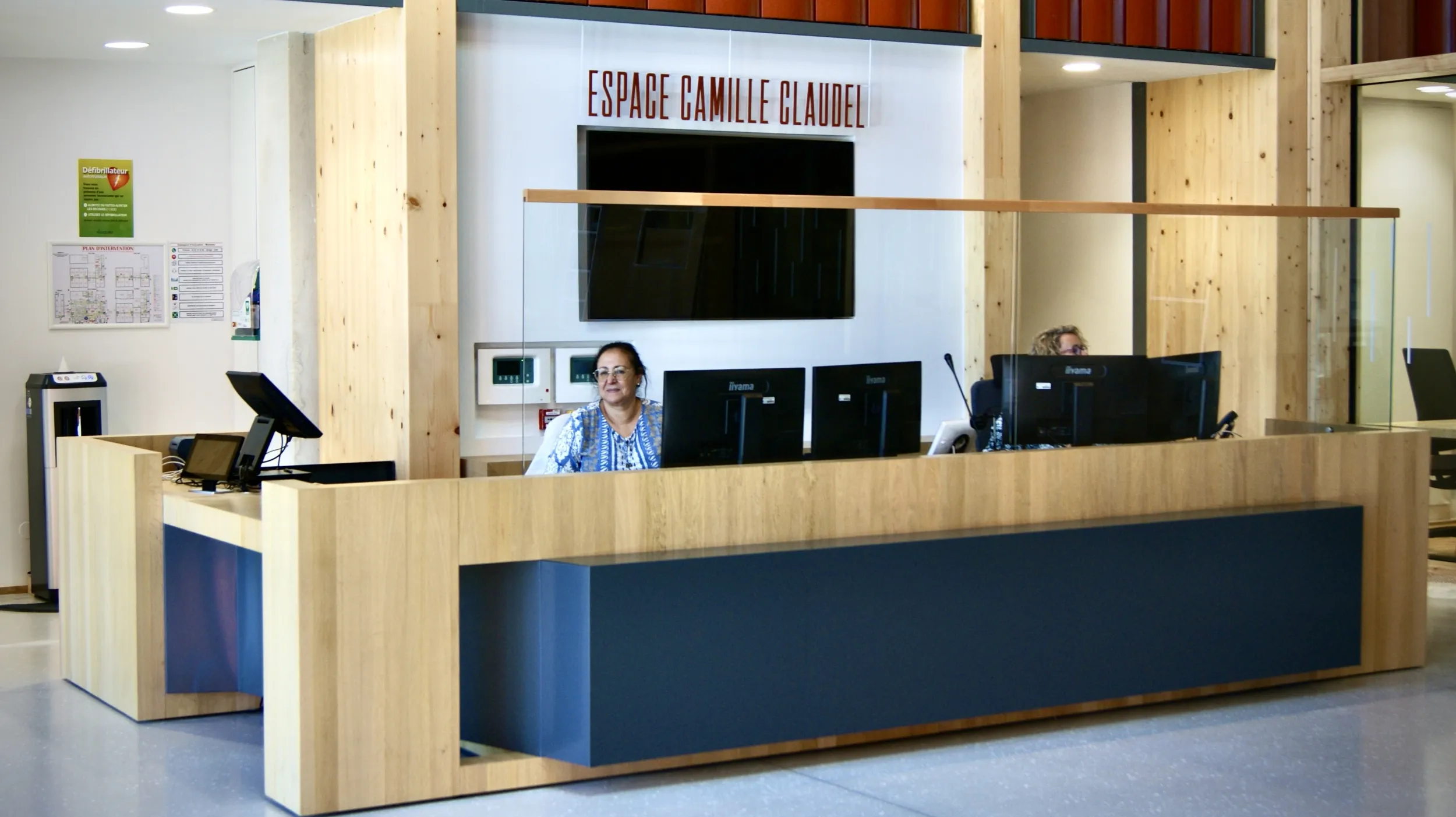 Derrière une banque d'accueil de bois, de métal, équipée d'écrans d'ordinateurs et surmontée d'une protection en plexiglas (?) , une dame souriante fixe l'objectif