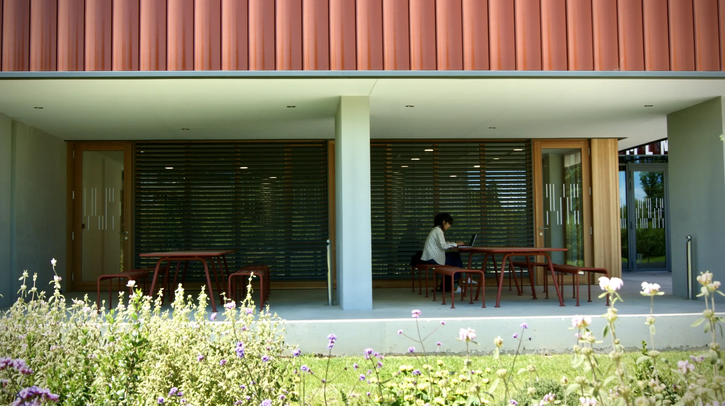 Vue sur deux arcades en béton surmontées de plaques tuilées ocrées, équipées de mobilier métallique d'extérieur, baies vitrées en arrière plan ; une personne assise sur l'un des bancs, travaille sur un ordinateur