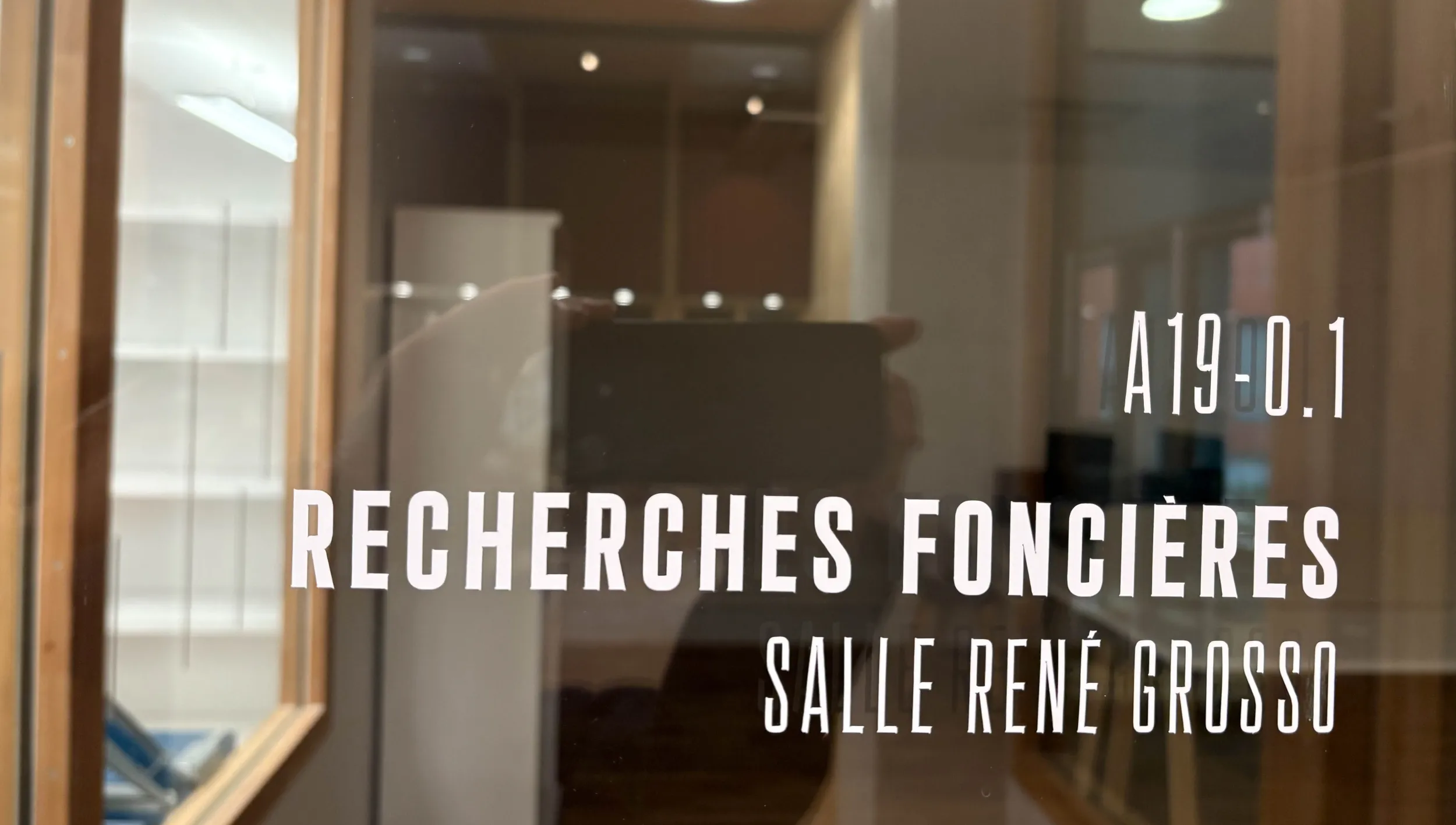 Signalétique en lettres blanches sur surface vitrée indiquant portant les mentions : A19.0.1 Recherches foncières, Salle René Grosso