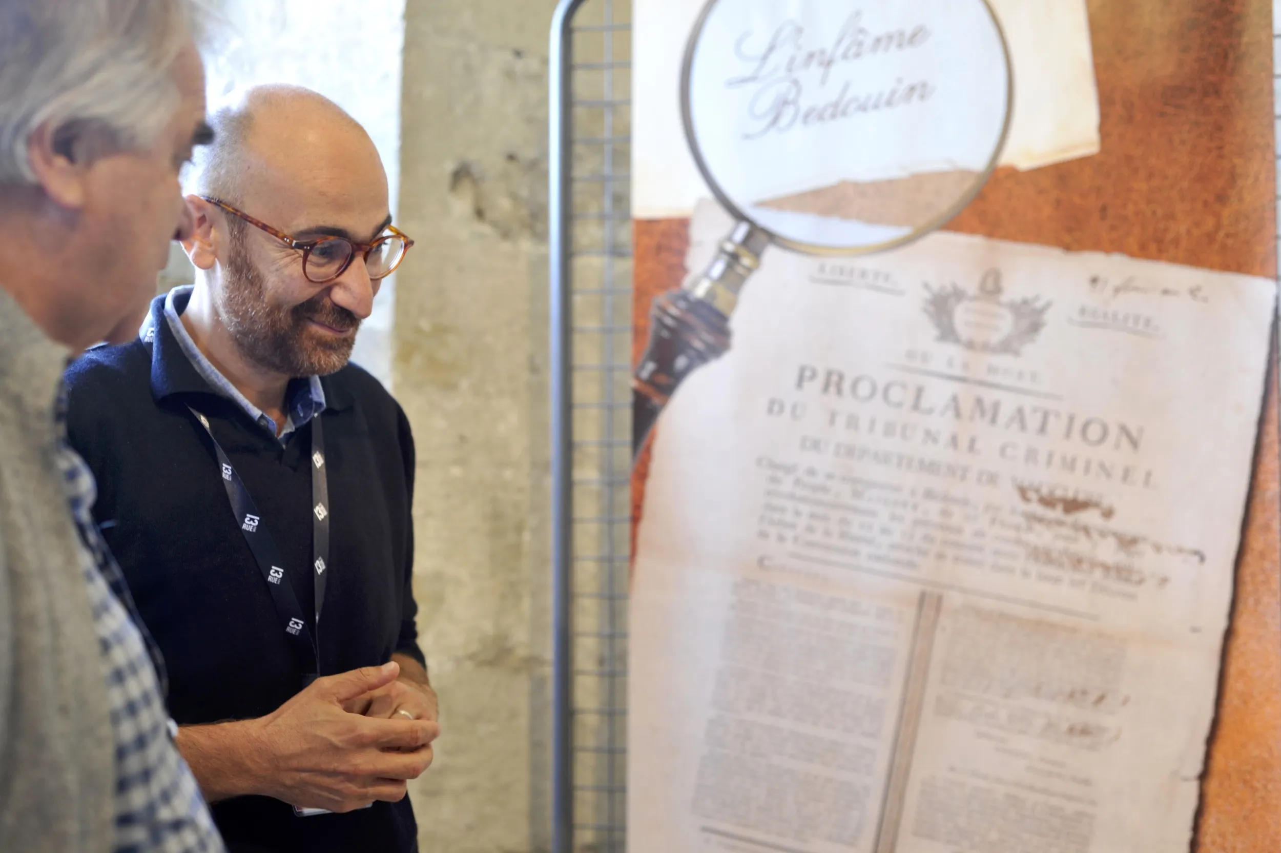 2 hommes devant un panneau d'exposition sur lequel figurent notamment "L'Infâme Bedouin" et "Proclammantion du tribunal criminel", le reste est flou.