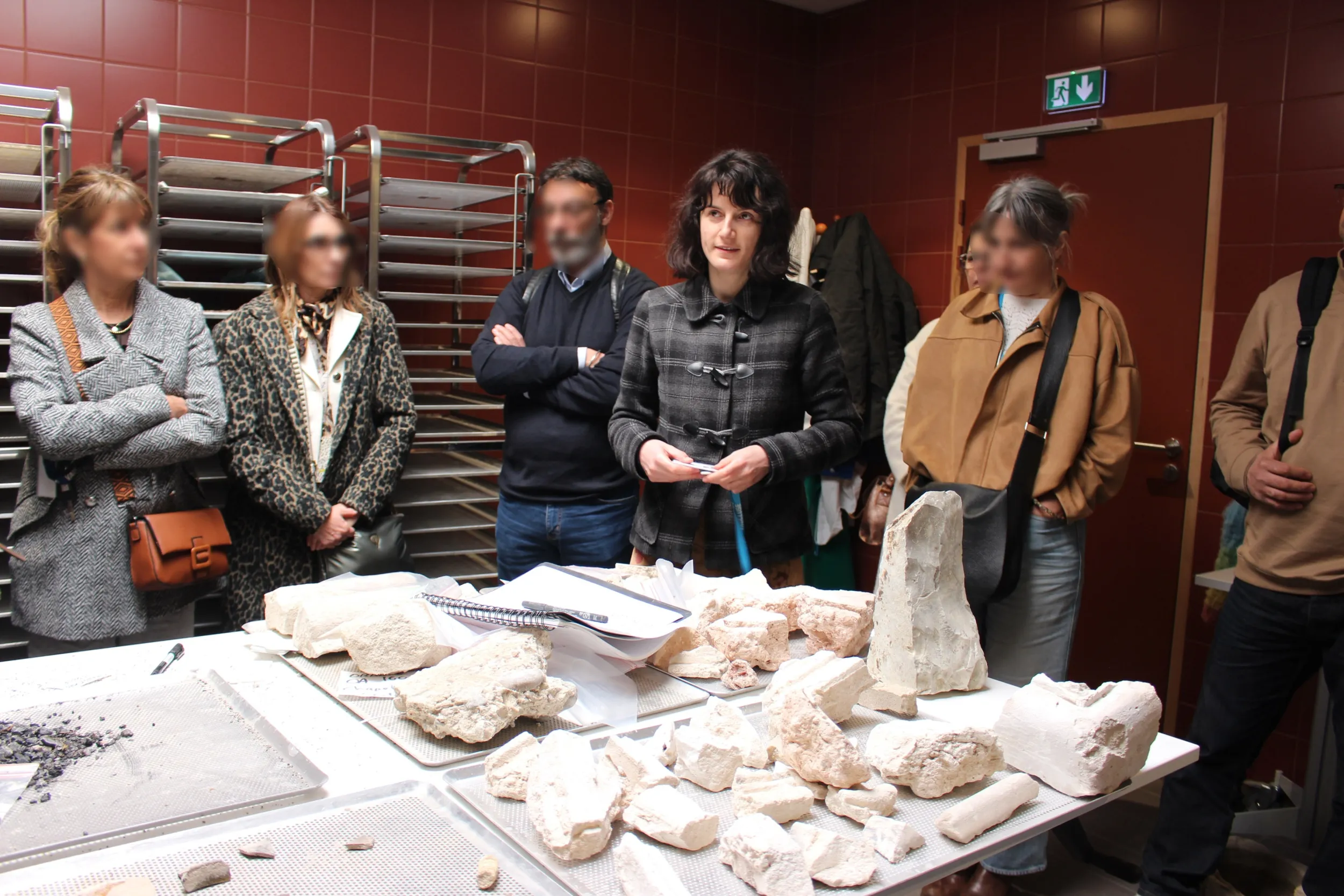 Une médiatrice entourée de 6 visiteurs présente des objets archéaologiques, à l'arrière des colonnes de séchage métalliques