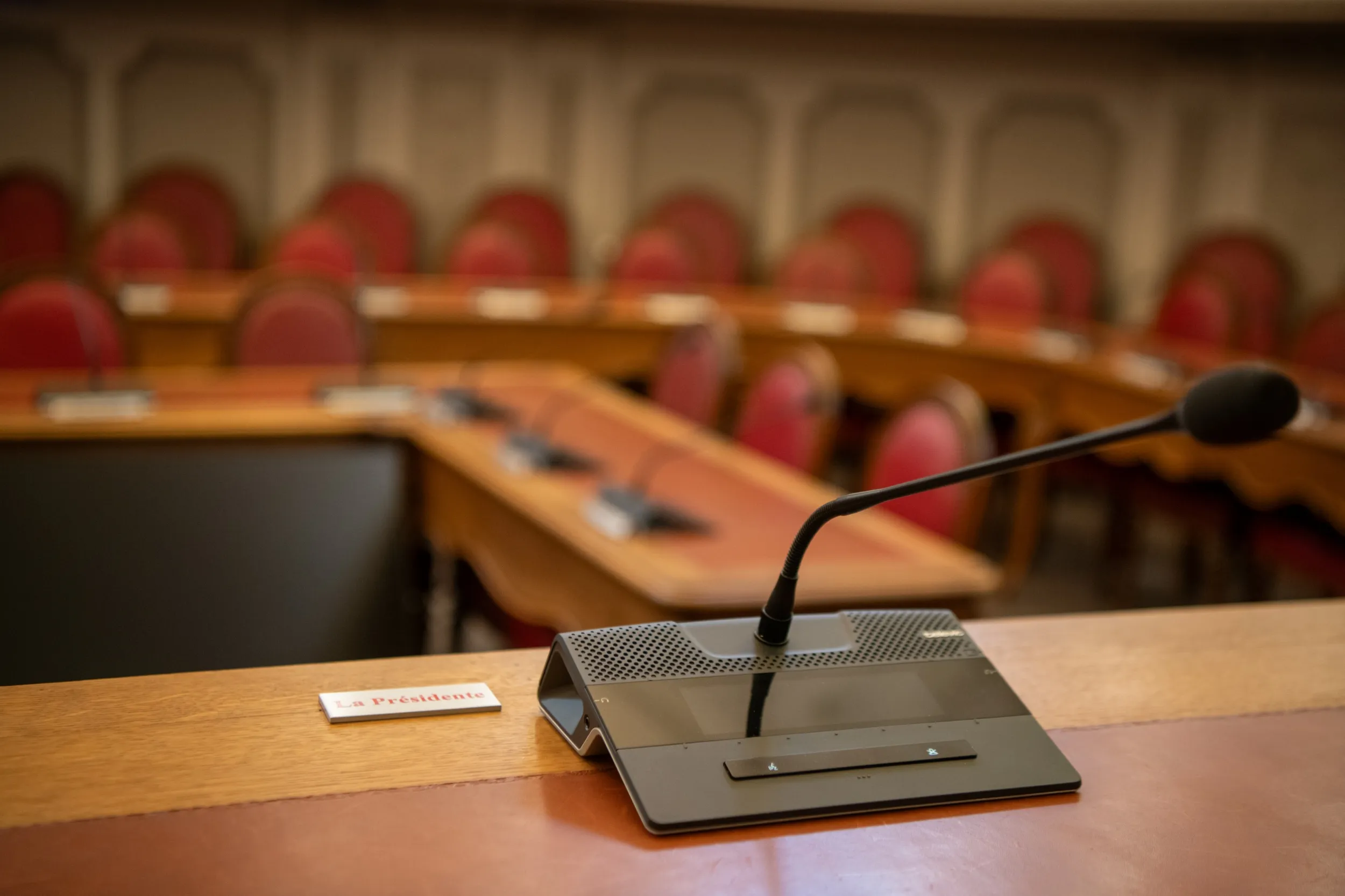 Vue depuis le siège de la présidence de séance, un micro au premier plan et au second plan, flouté, tables en U et fauteuils rouge