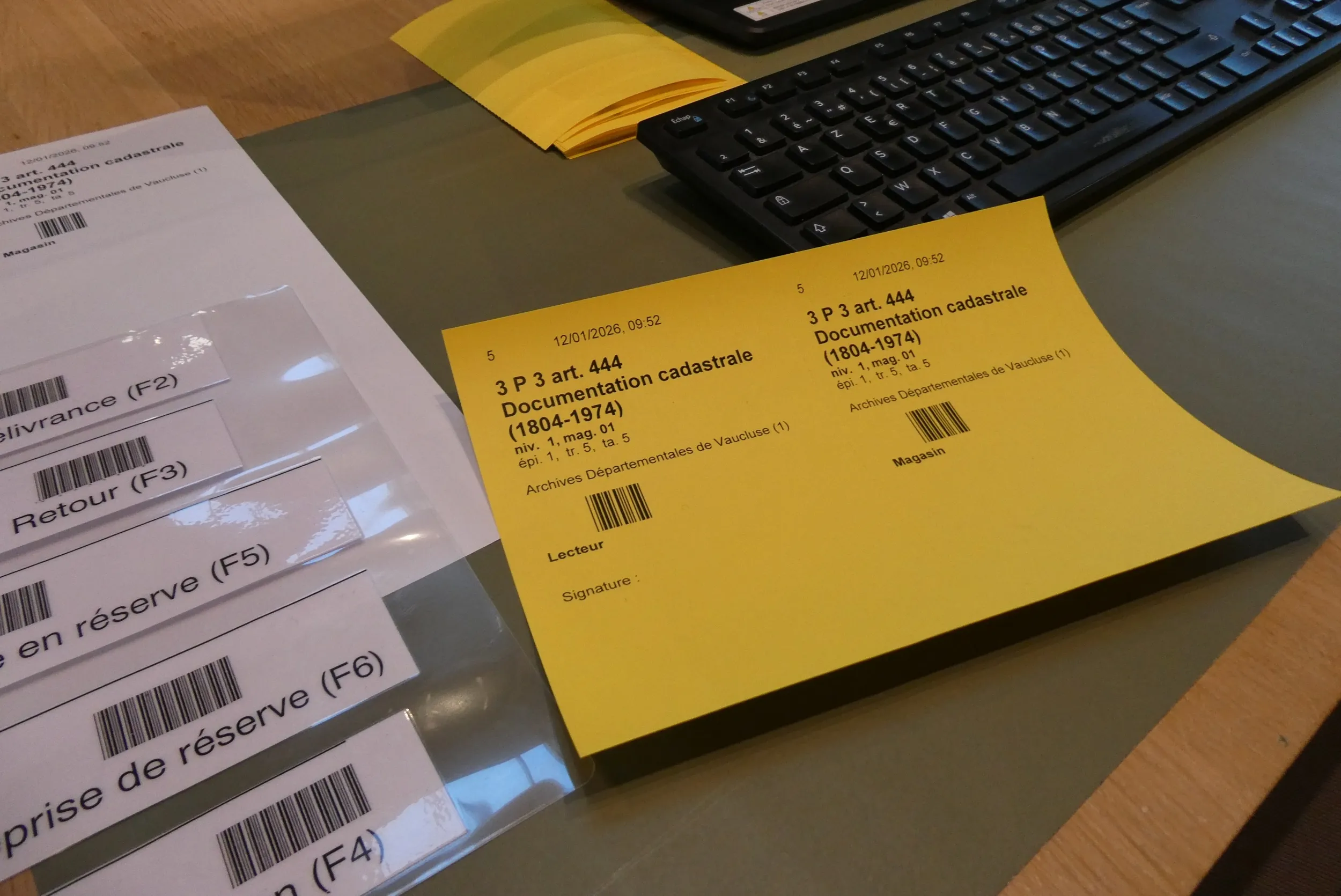 A côté d'un clavier d'ordinateur, fiche cartonnée jaune en deux parties sur laquelle figurent les mentions de : la date, les références du document, sa localisation, des codes barre et sur une fiche des partie un emplacement pour la signature.
