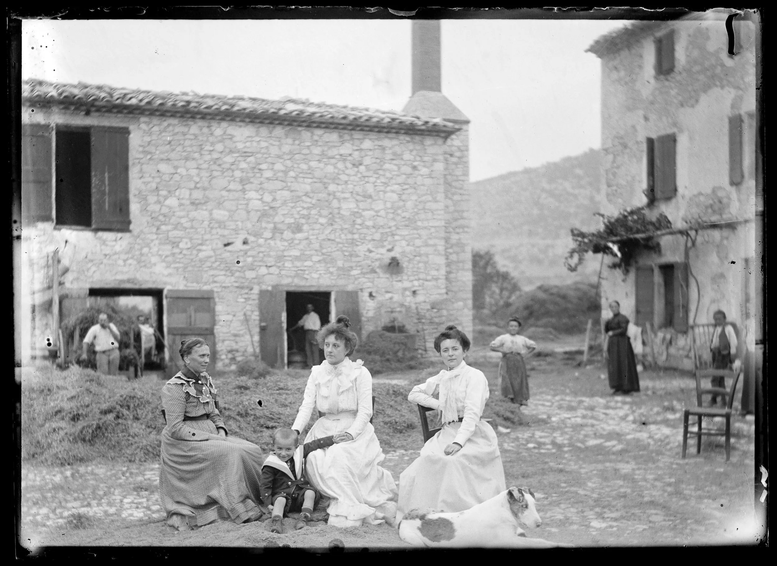 Carte postale en noir et blanc représentant : 3 femmes assises devant une distillerie, cinq autres personnes debout en arrière plan.