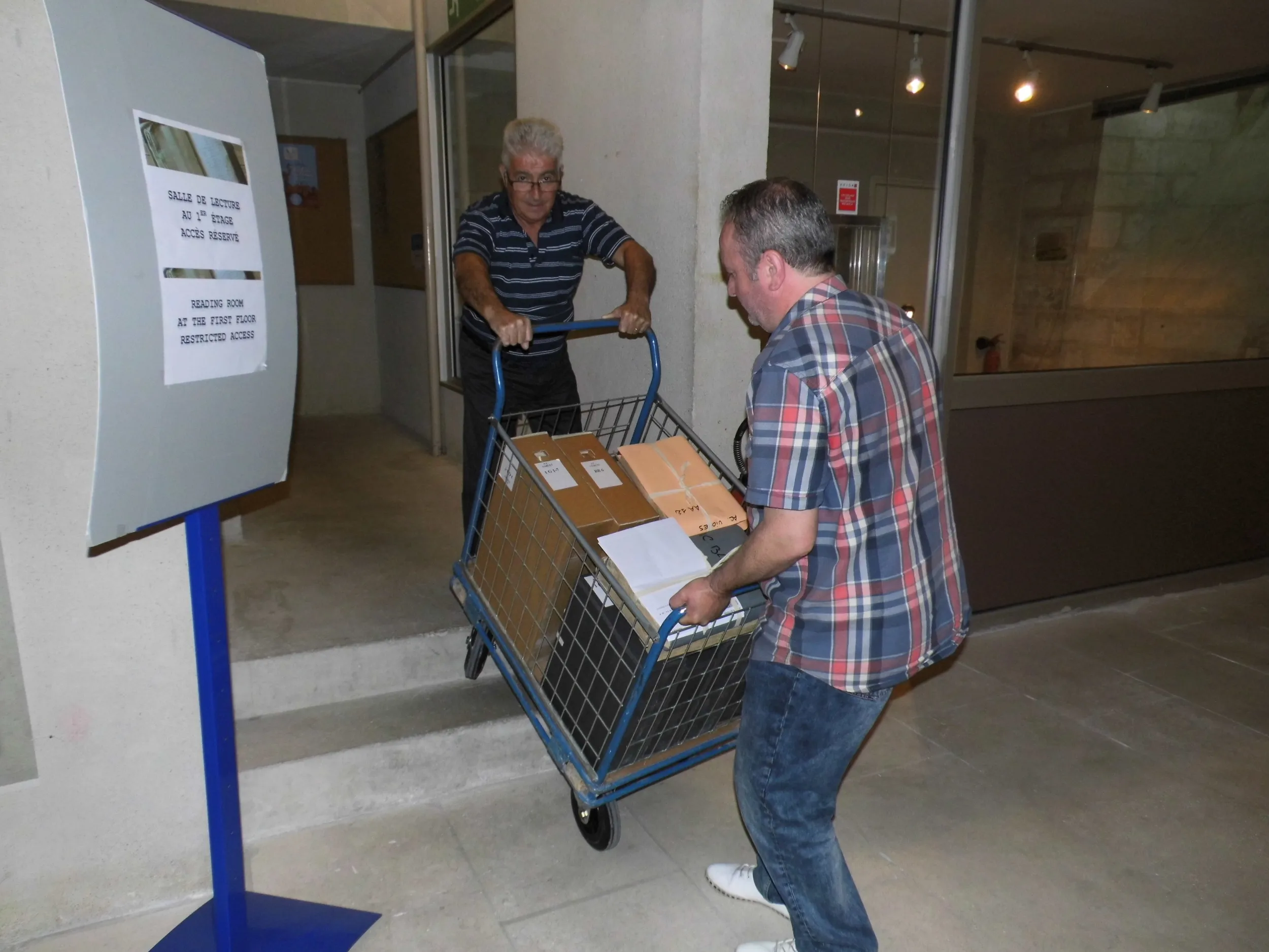 Deux hommes tentent de hisser un chariot rempli de cartons sur un escalier