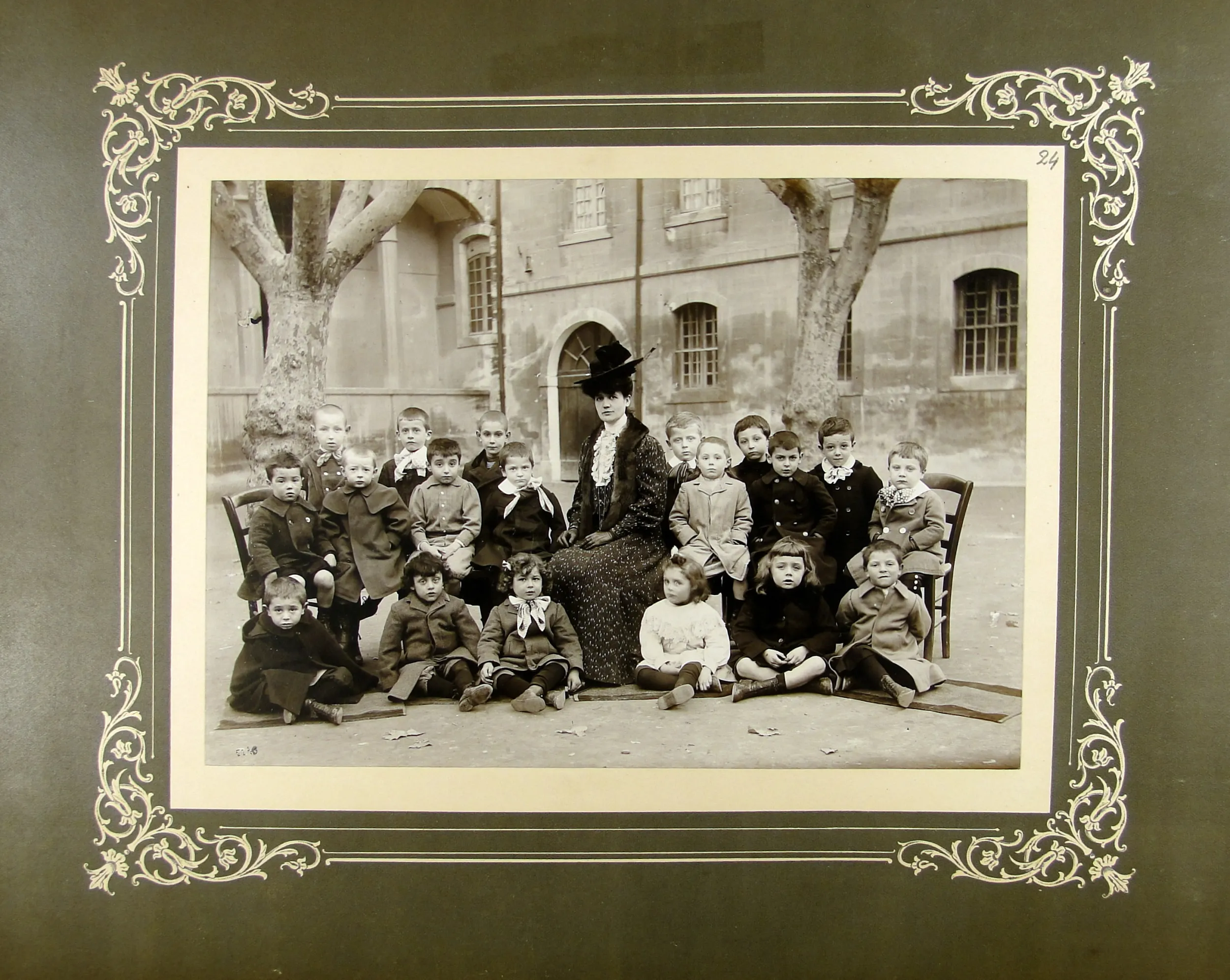 Photographie en noir et blanc de la fin du XIXe siècle montrant une institrice chapeautée entourée d'enfants posant pour la photo de classe dans une cour d'école.