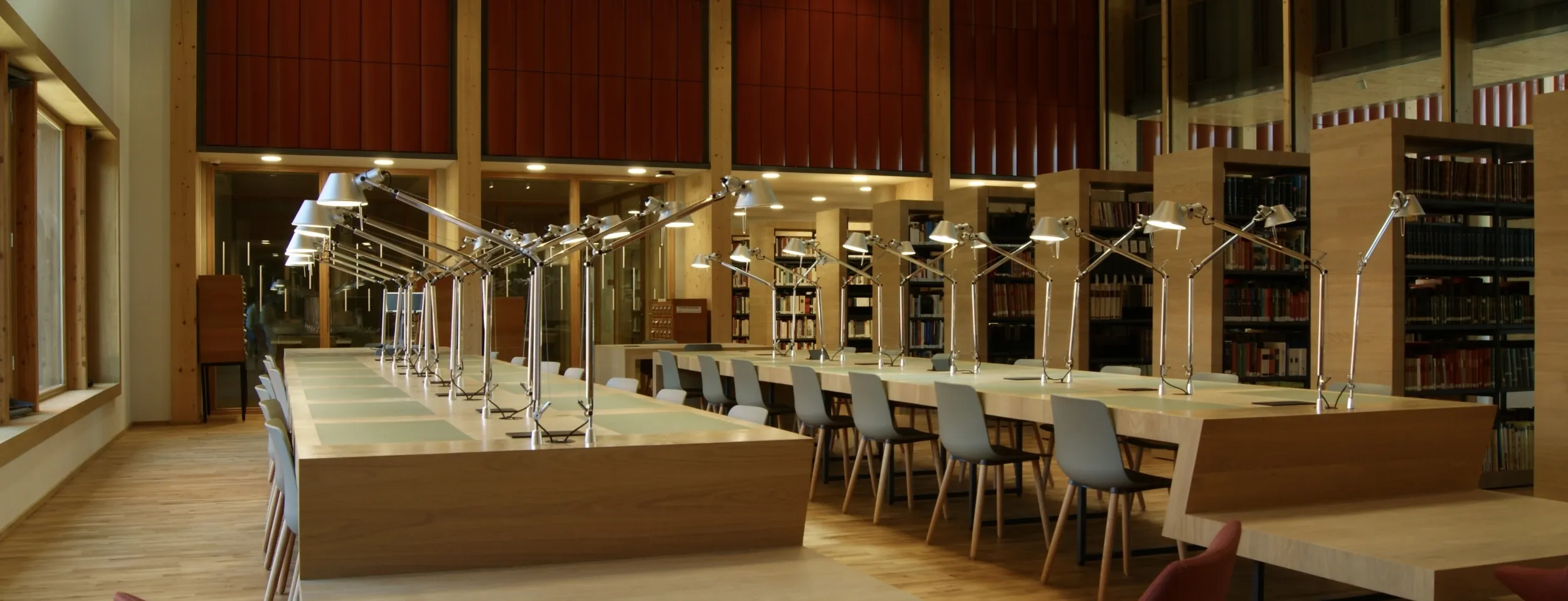 Vue en profondeur sur une salle de consultation comportant deux longues tables de travail en bois, surmontées de hautes lampes orientables ; à gauche, une baie vitrée, à droite, une bibliothèque