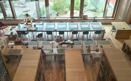 Dans une grande salle équipée de mobilier bois : rayonnages de bibliothèque, 2 grandes tables de travail, baies vitrées en fond, des chercheurs consultent des documents