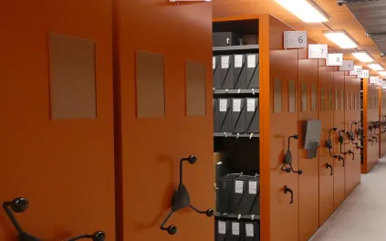 Vue sur des compactus métalliques de couleur orange ; sur les tablettes, des boîtes en carton de couleur noires sur lesquelles figurent des références