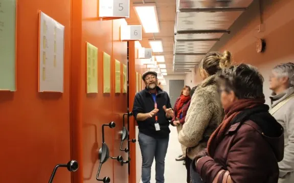 Dans un magasin d'archives modernes aux rayonnages métalliques de couleurs orange et grise, un archiviste guide des visiteurs